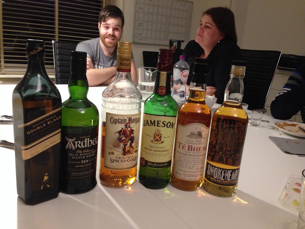 A row of various liqour bottles on a table. In the background, Tim Luigjes and Iacobien Riezebosch can be seen.