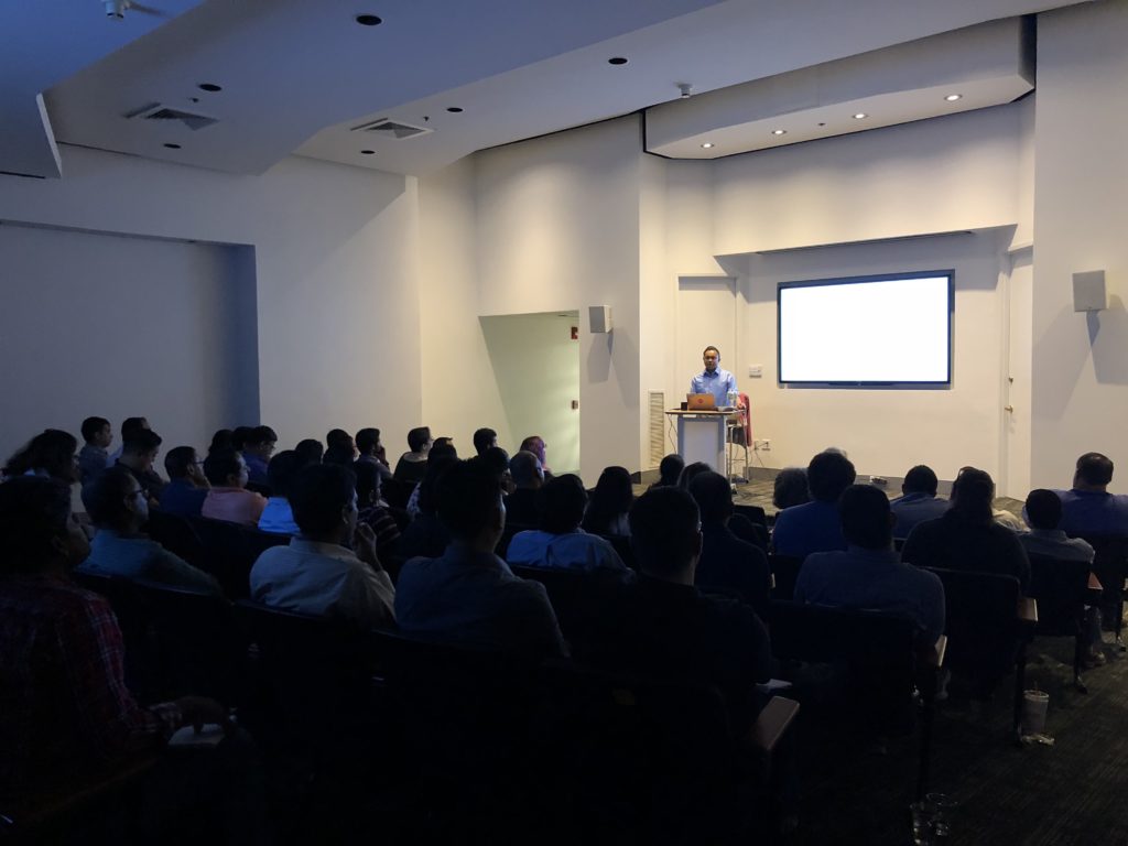 Single presenter on small stage, presenting to auditorium of people.